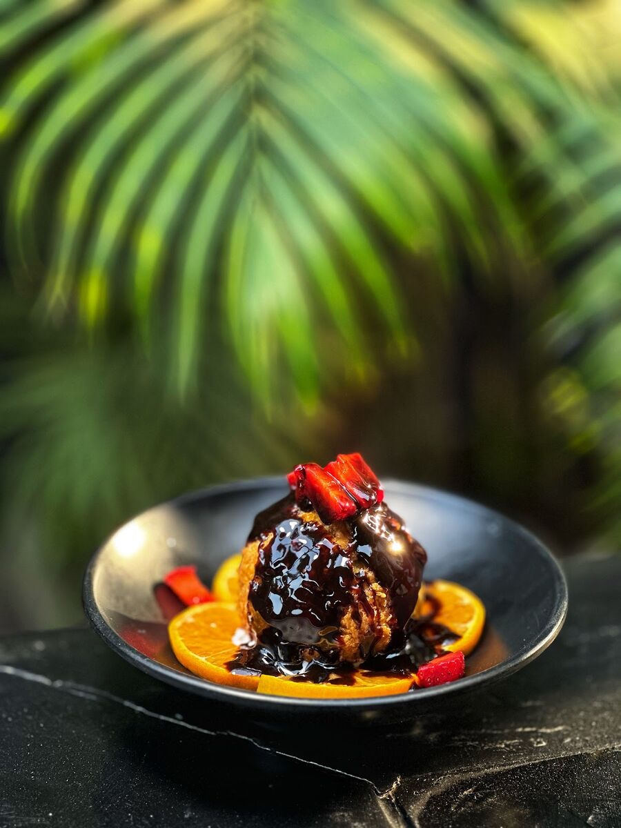 Round dessert covered in dark sauce with orange slices and red fruit on a black plate at EMC Kitchen